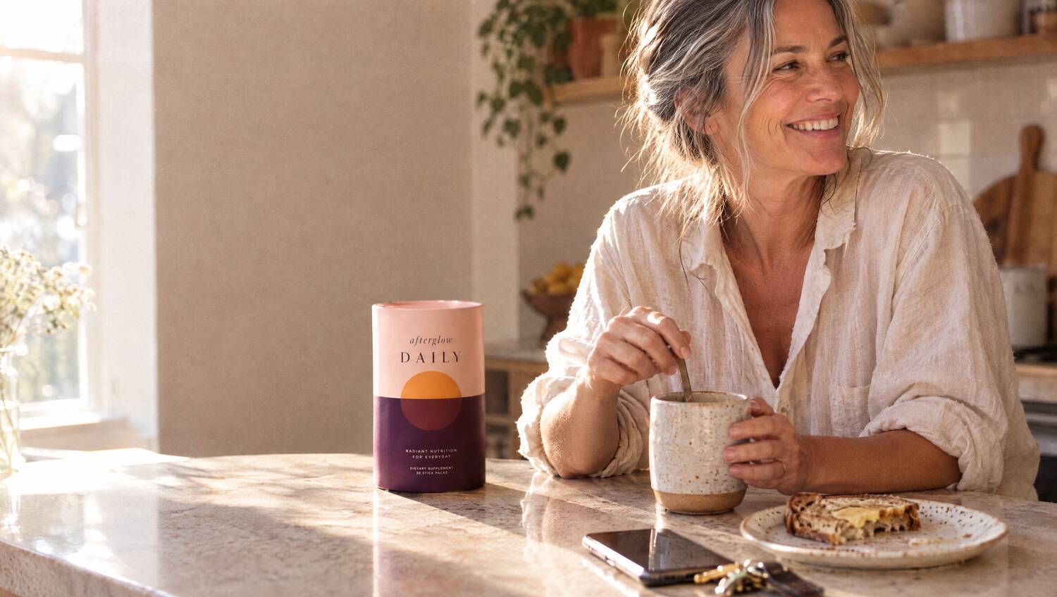 A smiling woman at her kitchen table with coffee and the Afterglow Daily tub
